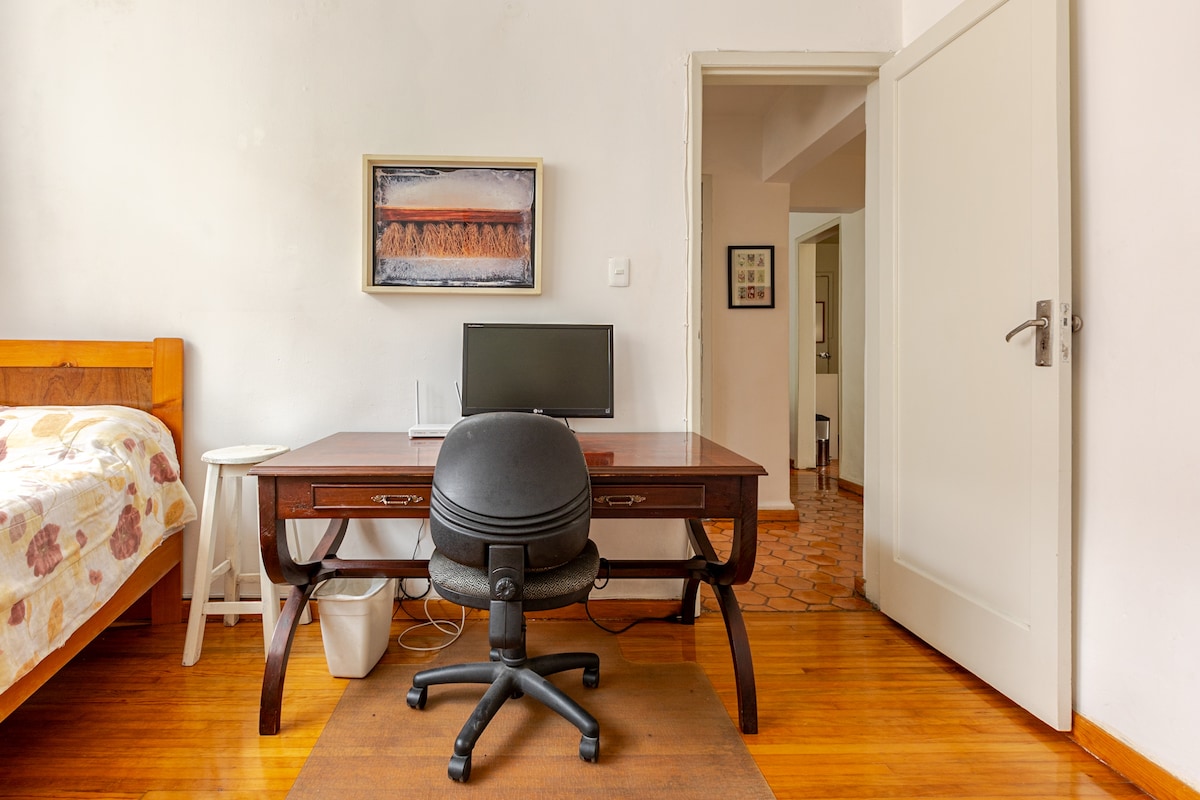 A workspace is set up with a wooden desk and an office chair. An HD television is positioned on the desk, and a door leads to another room. A small bedside table and a bed with a patterned blanket are visible, along with wooden flooring that enhances the space.