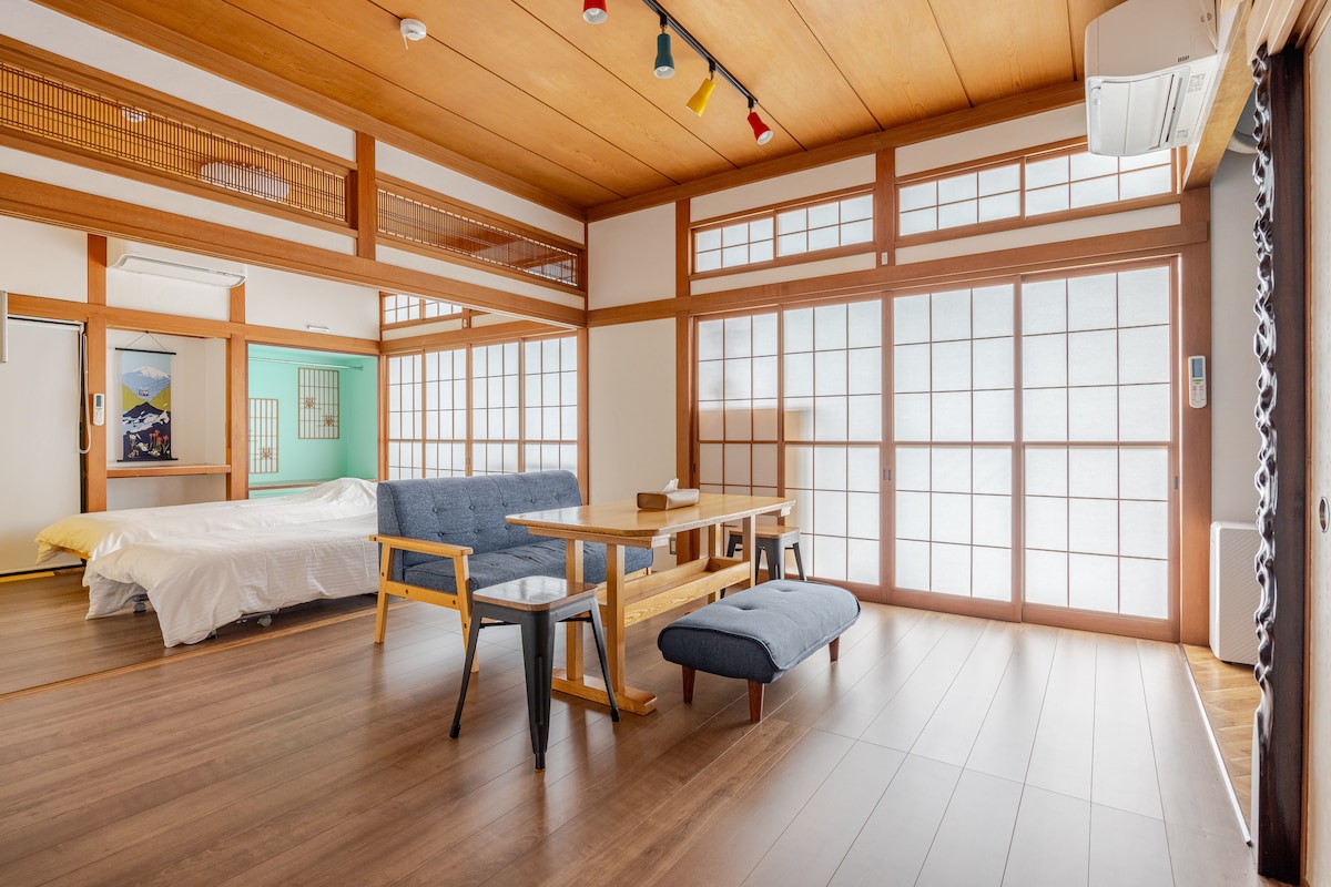 The spacious interior features traditional sliding shoji doors, allowing natural light to filter through. A dining table is paired with wooden chairs, complemented by a low seating area. A large bed is seen in the corner, with light hardwood flooring adding warmth to the space.