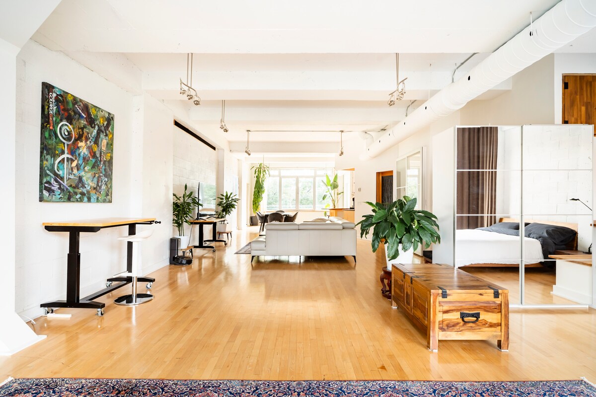 A spacious loft interior is revealed, featuring hardwood flooring, high ceilings, and an abundance of natural light from large windows. A mix of modern furnishings and greenery create a relaxed atmosphere. Distinct zones for living, dining, and sleeping are subtly defined, enhancing the open layout.