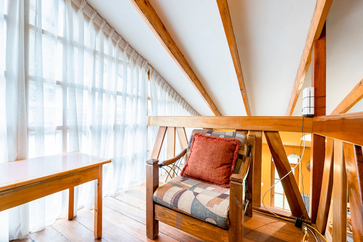 A cozy loft space features wooden beams and large windows draped with sheer curtains, allowing natural light to fill the area. A comfortable chair with a patterned cushion is placed next to a simple wooden table, creating a relaxed atmosphere.
