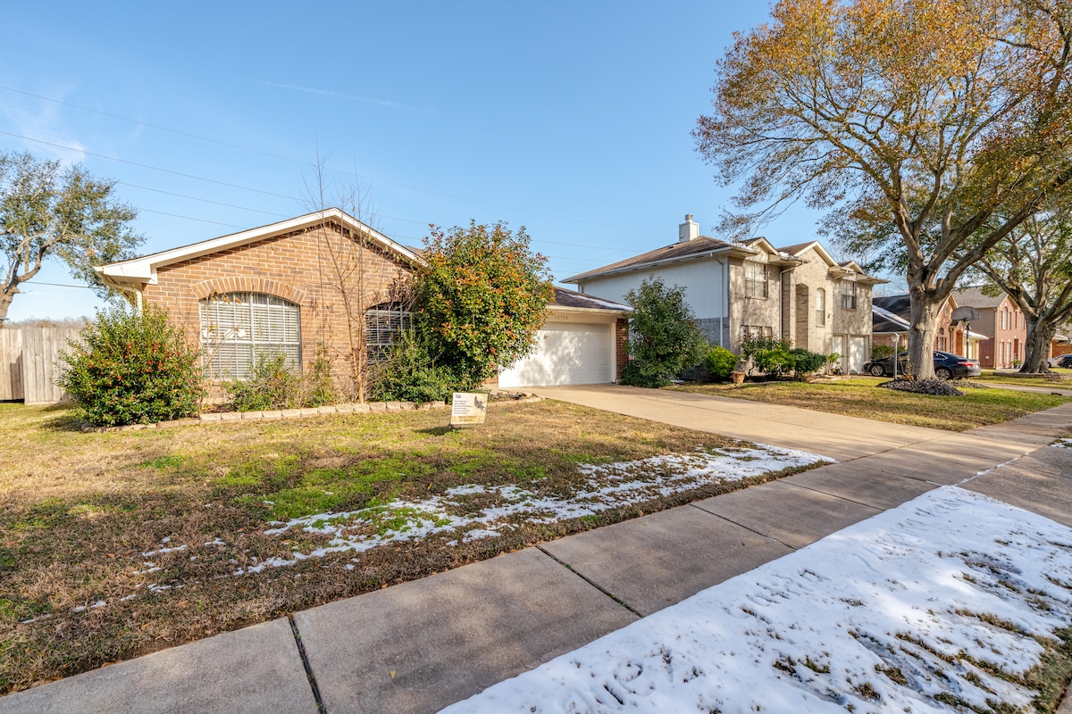 The image captures a single-story home with a brick facade and a textured driveway. Lush greenery surrounds the property, while patches of snow are visible on the sidewalk. Nearby houses and a clear blue sky complete the serene suburban atmosphere.