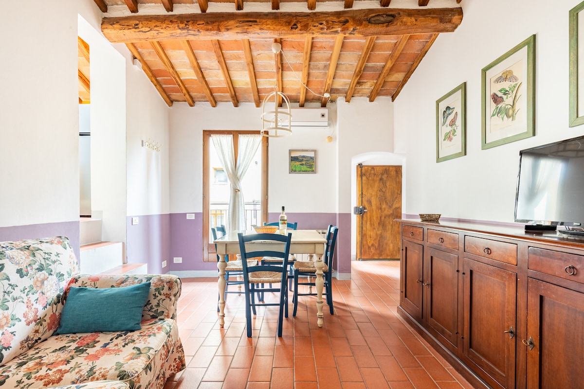 The living area features exposed wooden beams and terracotta flooring. A floral-patterned sofa complements a wooden dining table surrounded by blue and white chairs. Natural light enters through the window adorned with sheer curtains, enhancing the cozy atmosphere of the space.