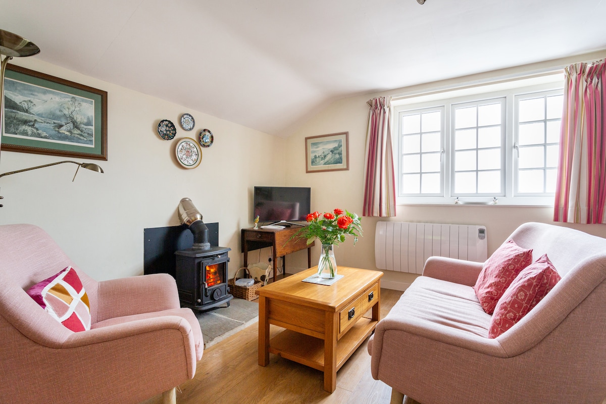 A cozy seating area features two pink armchairs and a wooden coffee table adorned with fresh flowers. A woodburning stove adds warmth, while a small television sits on a side table. Natural light filters through the multiple-paned windows dressed with light curtains.