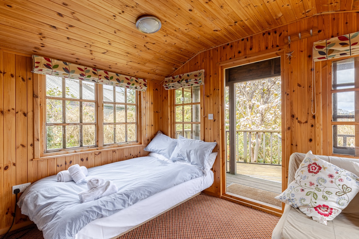 A cozy interior features wooden paneling and large windows allowing ample natural light. A spacious bed with white bedding is complemented by a single chair bed. A door leads to a deck area, providing access to outdoor views.
