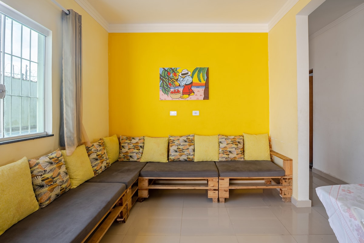 A cozy seating area is arranged with pallet sofas covered in soft cushions, featuring a bright yellow wall adorned with a colorful artwork. Large windows allow natural light to fill the space, enhancing the welcoming environment.