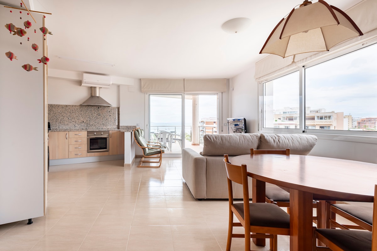 An open living space features a modern kitchen with a sleek range hood, complemented by a dining area with wooden chairs. A comfortable sofa is positioned within an environment filled with natural light, offering views through large windows that overlook the terrace and the beach.