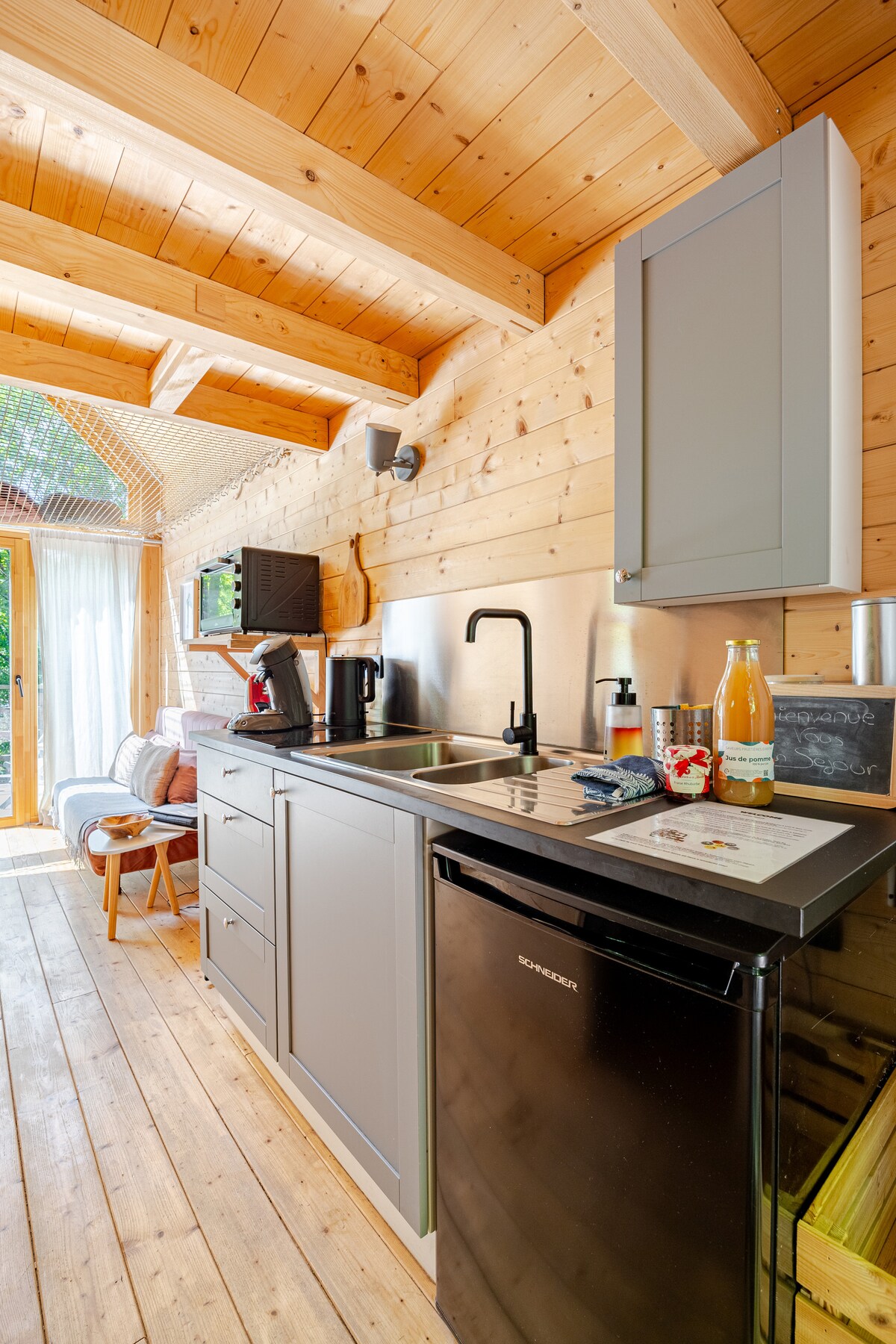 A compact kitchenette features wooden walls and ceiling beams, with modern appliances including a coffee machine and fridge. Natural light enters through a nearby window, illuminating the space. A small table is set nearby, highlighting the functional and inviting layout.