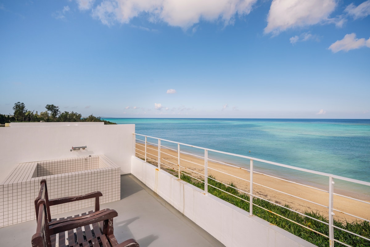 A private rooftop terrace features a scenic ocean view overlooking a sandy beach. A tiled hot tub is positioned near the edge, offering a relaxing spot. Wooden seating is provided, with the expansive seascape serving as a serene backdrop against a clear blue sky.