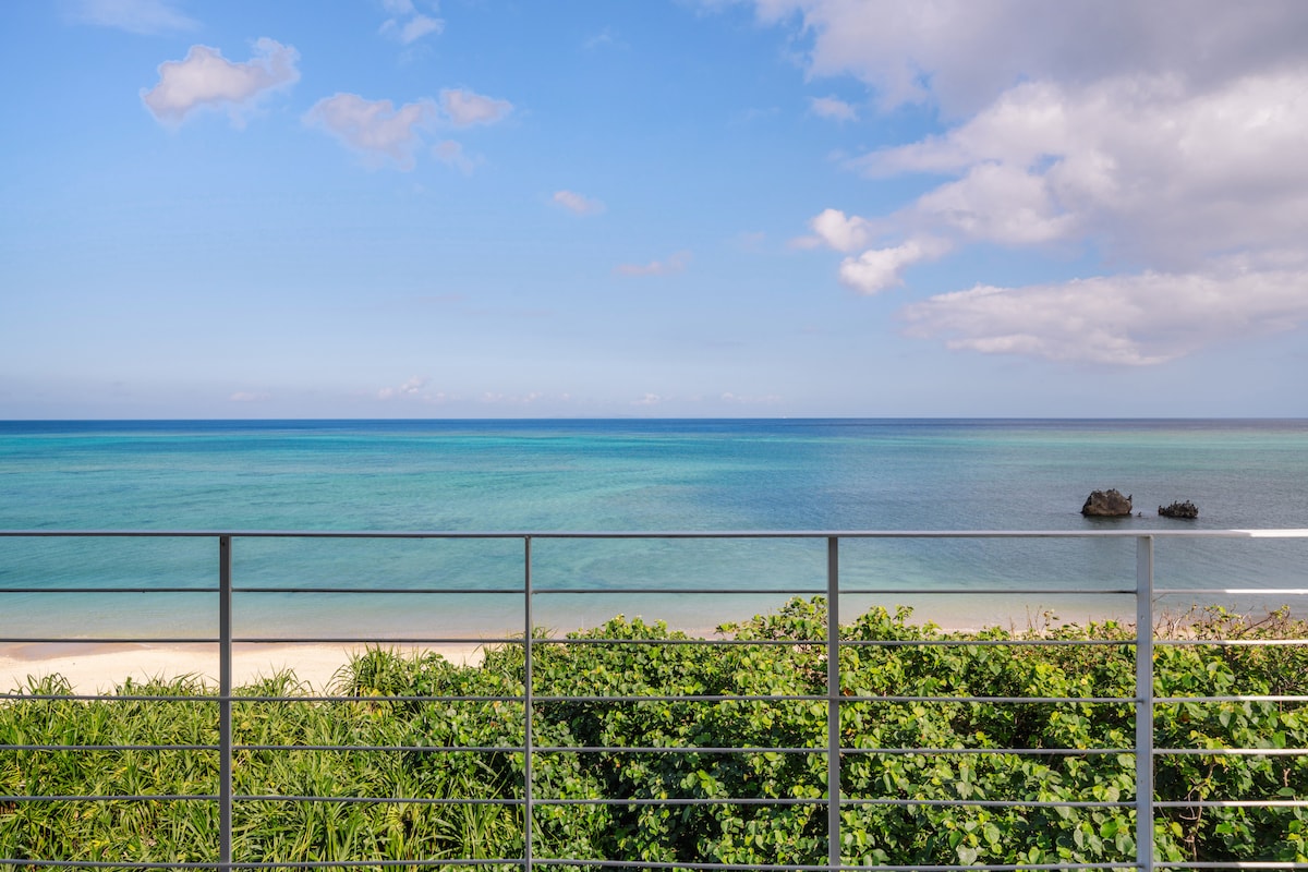 A view of the serene ocean is captured from a balcony railing, showcasing vibrant blue waters meeting a sandy beach. Lush greenery frames the scene, and distant rock formations are visible on the horizon, enhancing the tranquil coastal setting.