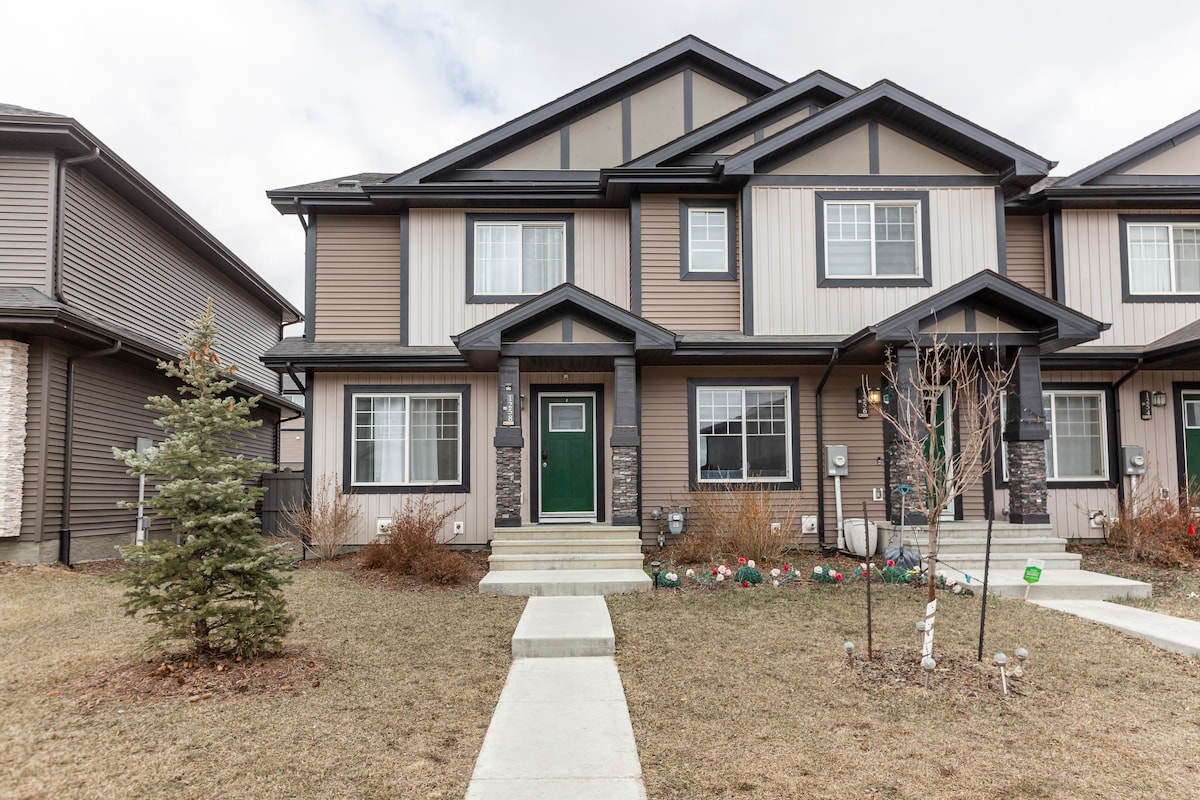 The front view of a two-story townhouse displays a well-maintained exterior, featuring a combination of earthy tones and stone accents. The entrance includes a green door, with windows flanking on either side. A neatly trimmed lawn and concrete walkway lead to the entrance.