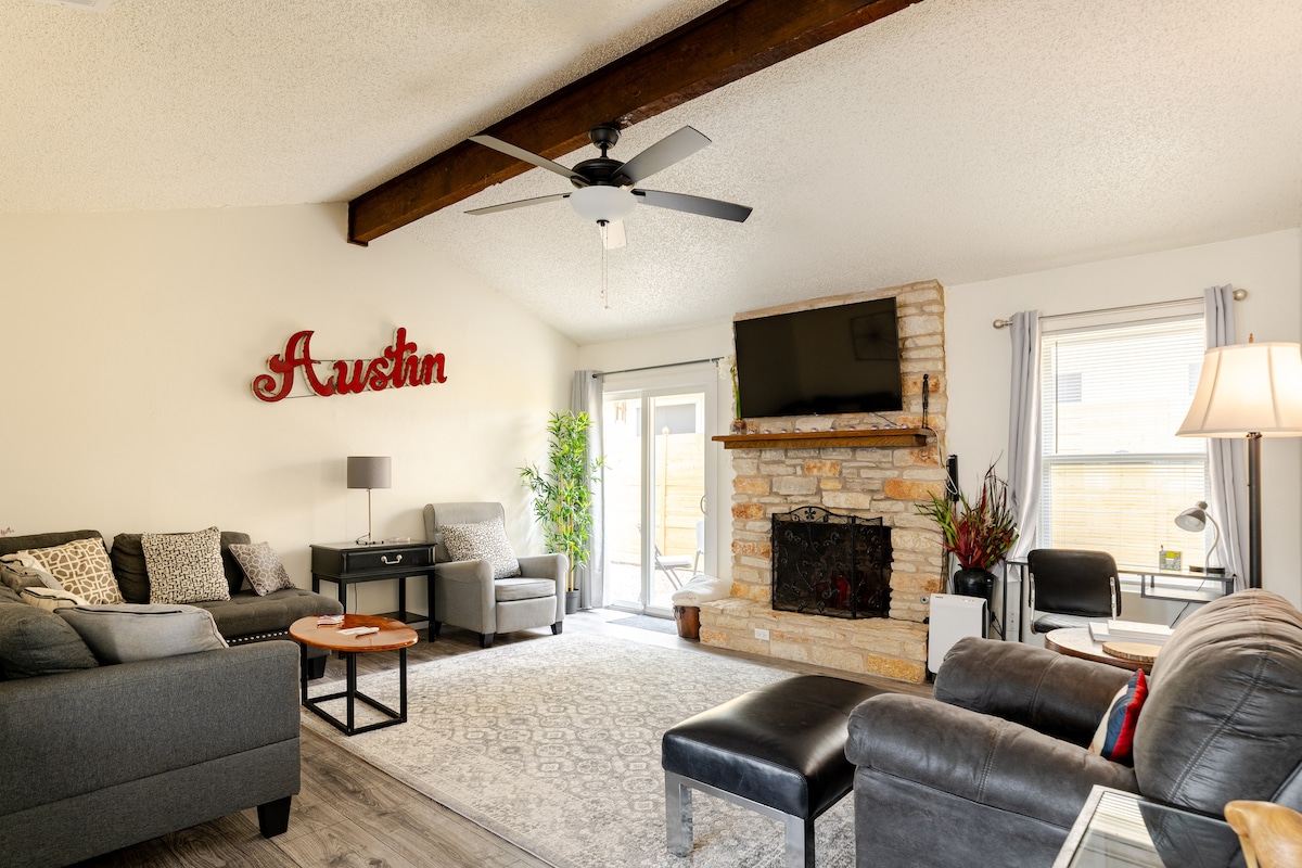 A spacious living area is highlighted by wood beams and a stone fireplace, featuring a comfortable grey sofa and matching armchair. Natural light filters through large windows, creating a welcoming environment. A television is mounted above the fireplace, and a small table is positioned nearby.