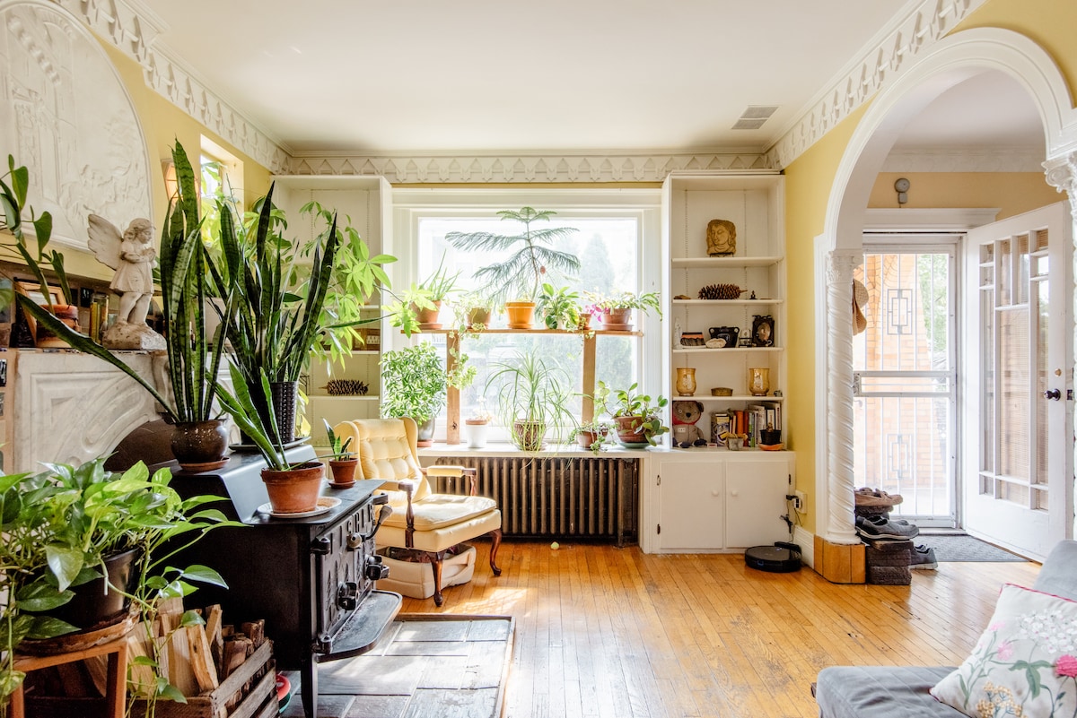 A bright living room features large windows adorned with potted plants, providing a view of greenery. A vintage wood stove sits beside a cozy armchair. Shelves display various decorative items, contributing to the warm and inviting atmosphere of the space.