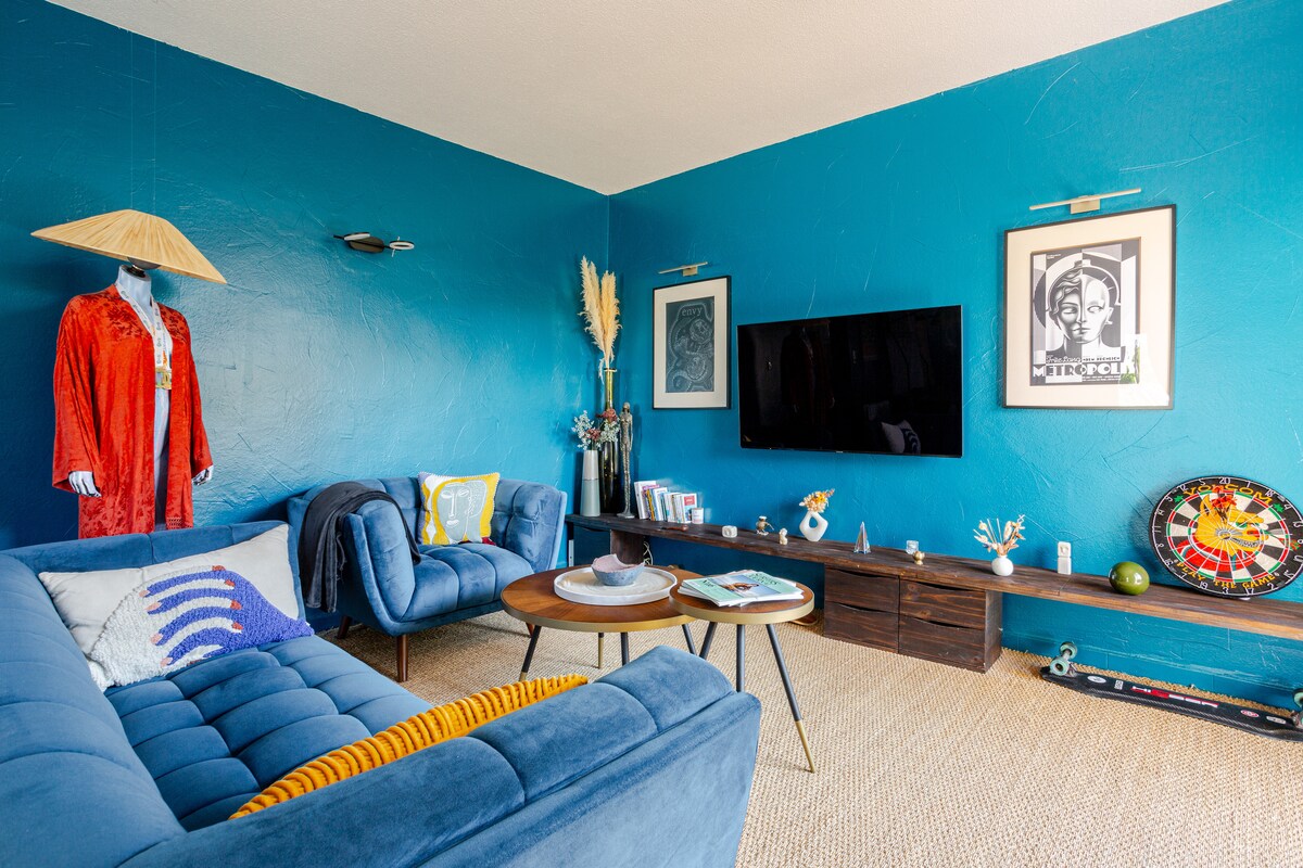 A living room features a deep blue accent wall, complemented by a light-coloured carpet. A modern television is mounted on the wall, with a wooden media unit below. Two comfortable couches provide seating, adorned with decorative pillows. An artwork and potted plants add character.