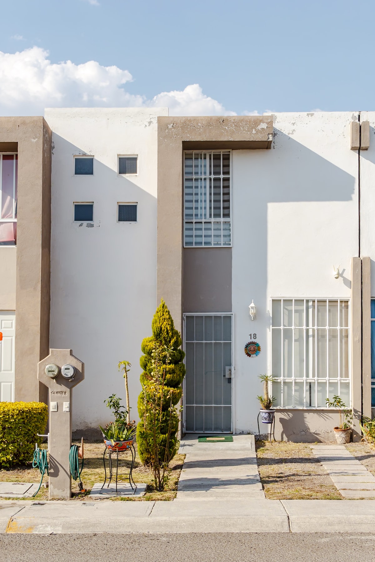 The exterior of a modern home is displayed, featuring a clean white façade with grey accents. A small garden with shrubs is positioned in front, alongside a decorative meter post. A tall, narrow window and a gate invite guests into the residence.