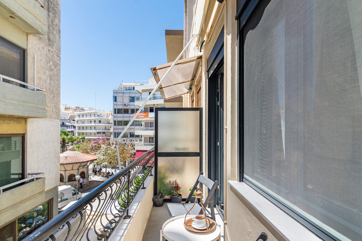 A balcony overlooks a bustling street, featuring a small table and two chairs. Sunlight brightens the space, and a potted plant adds a touch of greenery. The balcony's glass panels provide privacy while maintaining the connection to the lively surroundings.