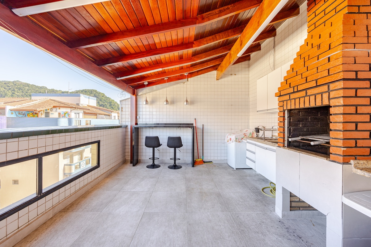 A spacious outdoor terrace features a traditional brick barbecue grill and two high-top stools positioned at a counter. The wood-beamed ceiling enhances the open-air design, while large windows offer views of the surrounding landscape.