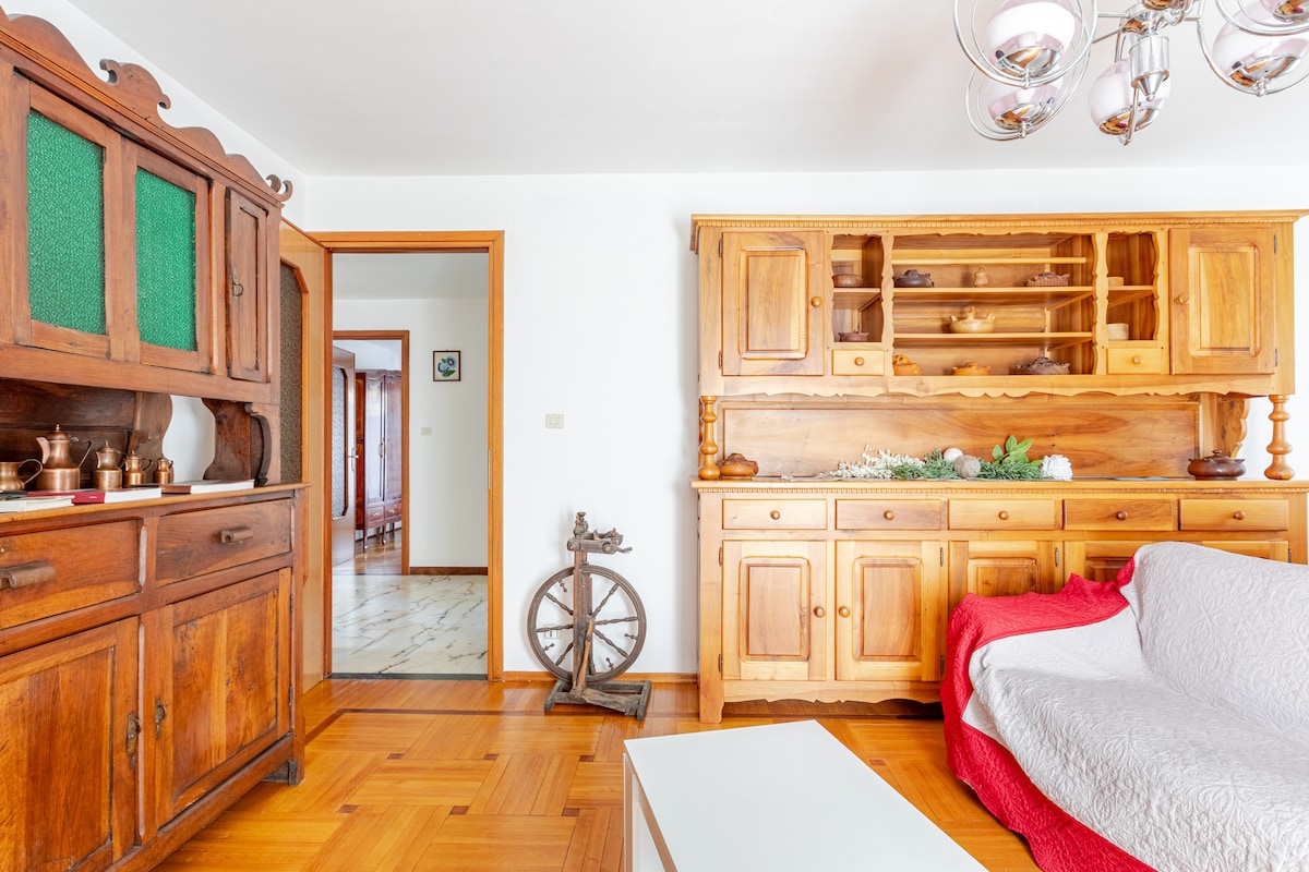 A warm living area features a wooden cabinet with green glass panels and decorative items, complemented by a cozy white sofa draped with a red blanket. The room includes a traditional spinning wheel and a white coffee table on a patterned floor, with doorways leading to additional spaces.