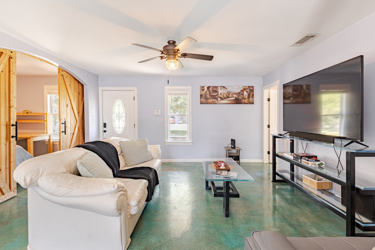 A spacious living area features a large sectional sofa with light upholstery and a coffee table positioned centrally. A television is mounted on a stand along one wall. Natural light enters through a front door and nearby window, illuminating the colorful floor.