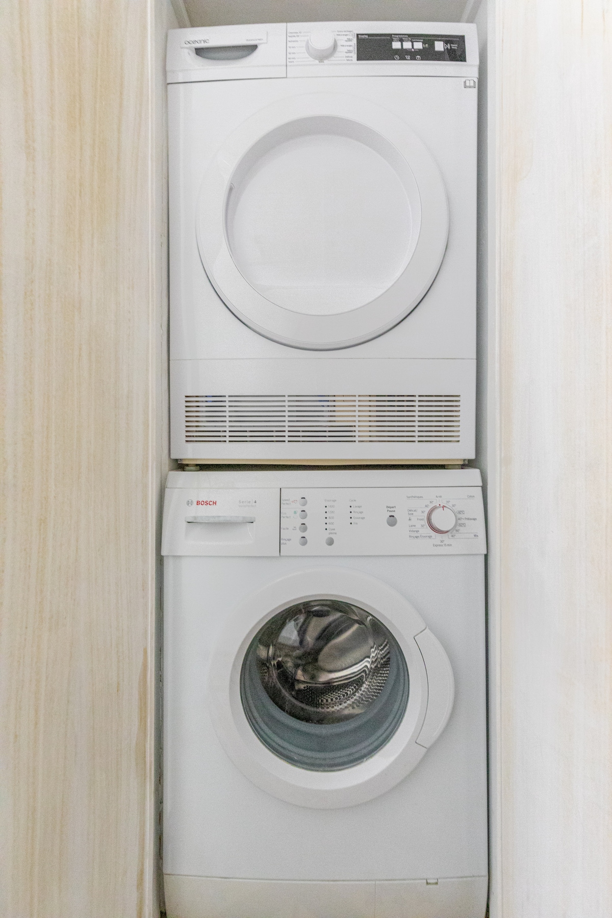 A stacked washer and dryer unit is positioned against a light-colored wall. The dryer is mounted on top, showcasing a round door and control panel, while the washing machine below features a transparent front door and its own control buttons.