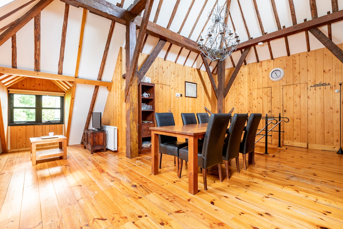 A spacious common area features a large wooden dining table surrounded by six black chairs. Exposed wooden beams and warm wooden walls create a rustic feel. A chandelier hangs from the ceiling, and a small coffee table is positioned nearby, enhancing the inviting ambiance.