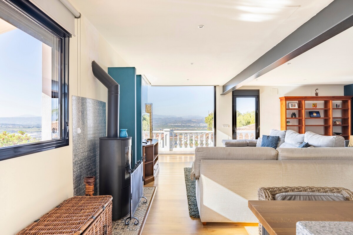 The living area features a comfortable sectional sofa, complemented by a natural wood coffee table. Large glass doors open up to a balcony, providing expansive views of the surrounding landscape. A modern wood stove adds a cozy element to the open space.