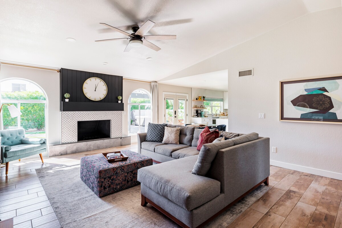 The living area features a spacious sectional sofa, a large coffee table, and a decorative rug. Natural light fills the room through large arched windows. A modern fan circulates air, while a wall clock and abstract art add visual interest. The kitchen is visible in the background.