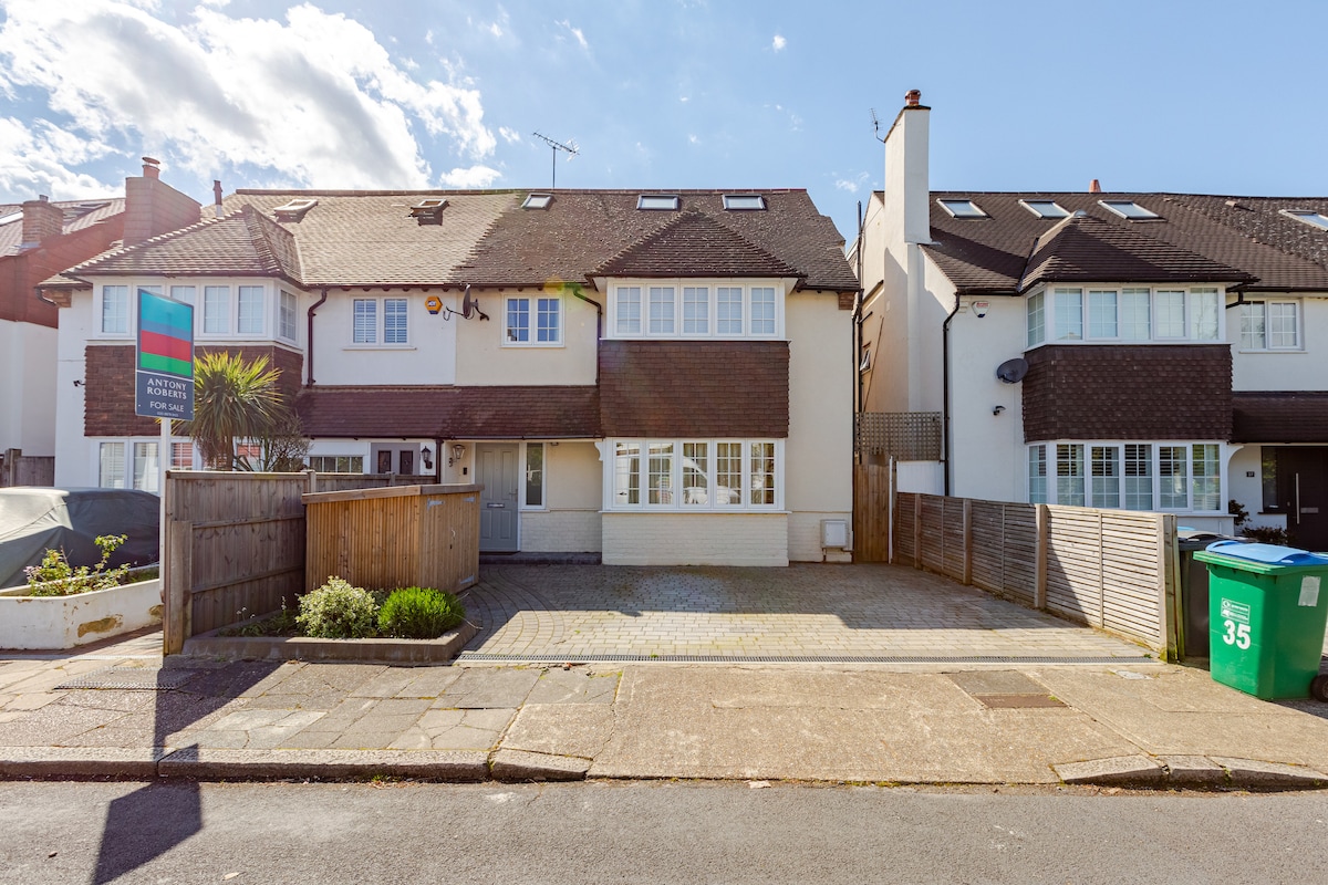 The exterior view of a spacious two-story home is presented, showcasing a well-maintained façade with multiple windows. A paved driveway allows for parking, and a low wooden fence surrounds the front garden, which features some greenery. The sky is clear with fluffy white clouds.