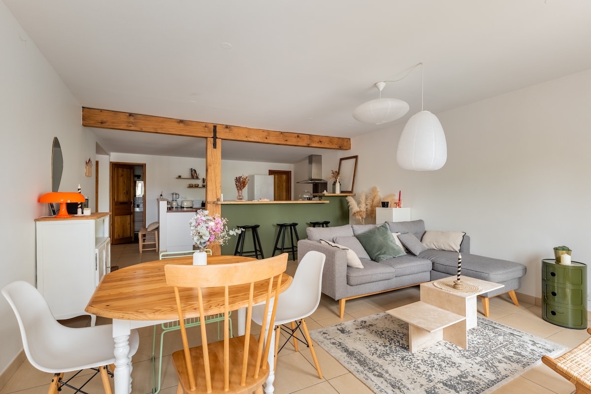 A modern living and dining area displays a blend of functional furniture. A gray sectional sofa is accompanied by a coffee table and a light-colored rug. A round dining table for four is set beside a kitchenette, while decorative accents, such as flowers and lighting fixtures, add character.