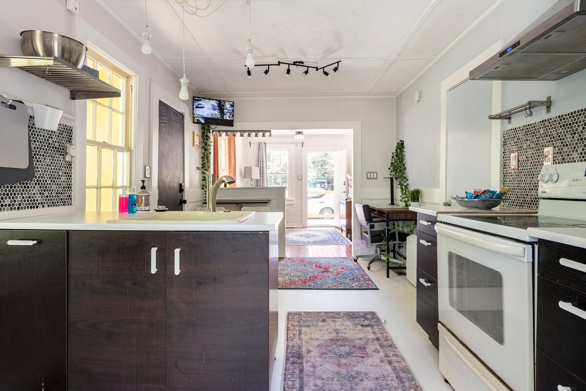 A well-equipped kitchen is showcased with modern dark cabinetry and a white countertop. A patterned rug adds a touch of color to the space. Natural light floods in through the windows, with a view of the living area and an outdoor pathway visible in the background.