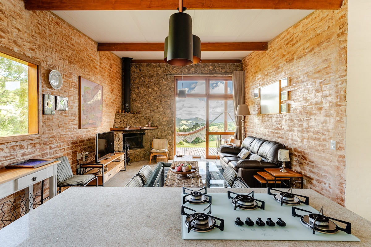 A modern living space features exposed brick walls and a cozy seating area with a dark leather couch. A stone fireplace is positioned next to large windows providing scenic views. A well-equipped kitchen showcases a cooktop on a light granite countertop, enhancing the functional layout.