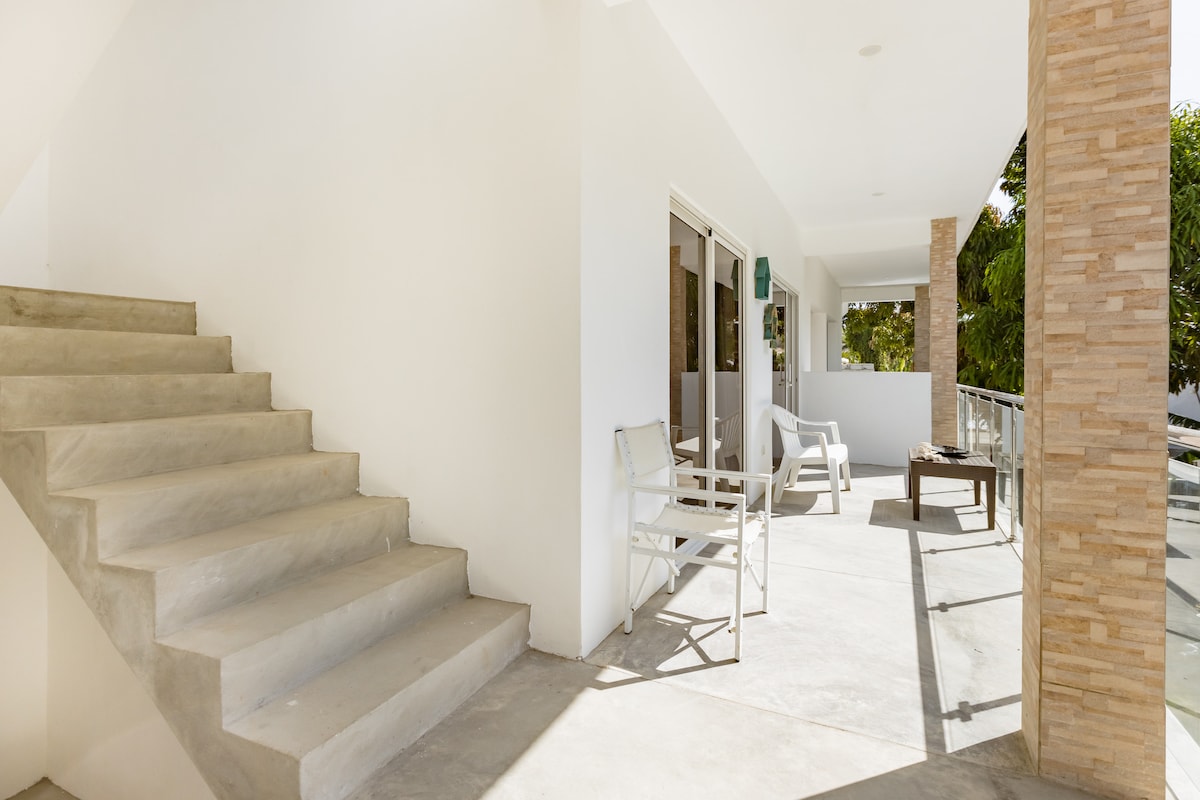 A spacious balcony is visible, featuring a seating area with white chairs and a small table. A flight of concrete stairs leads up to the balcony, which is framed by light-colored walls and natural stone columns. Abundant natural light fills the area.