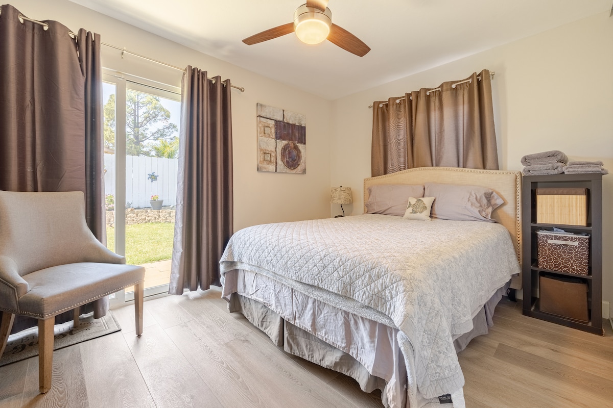 A well-appointed bedroom features a spacious bed with light bedding and decorative pillows. A comfortable chair is positioned beside a nightstand. Large windows with dark curtains allow natural light to enter, creating a bright atmosphere while offering views of the serene backyard.