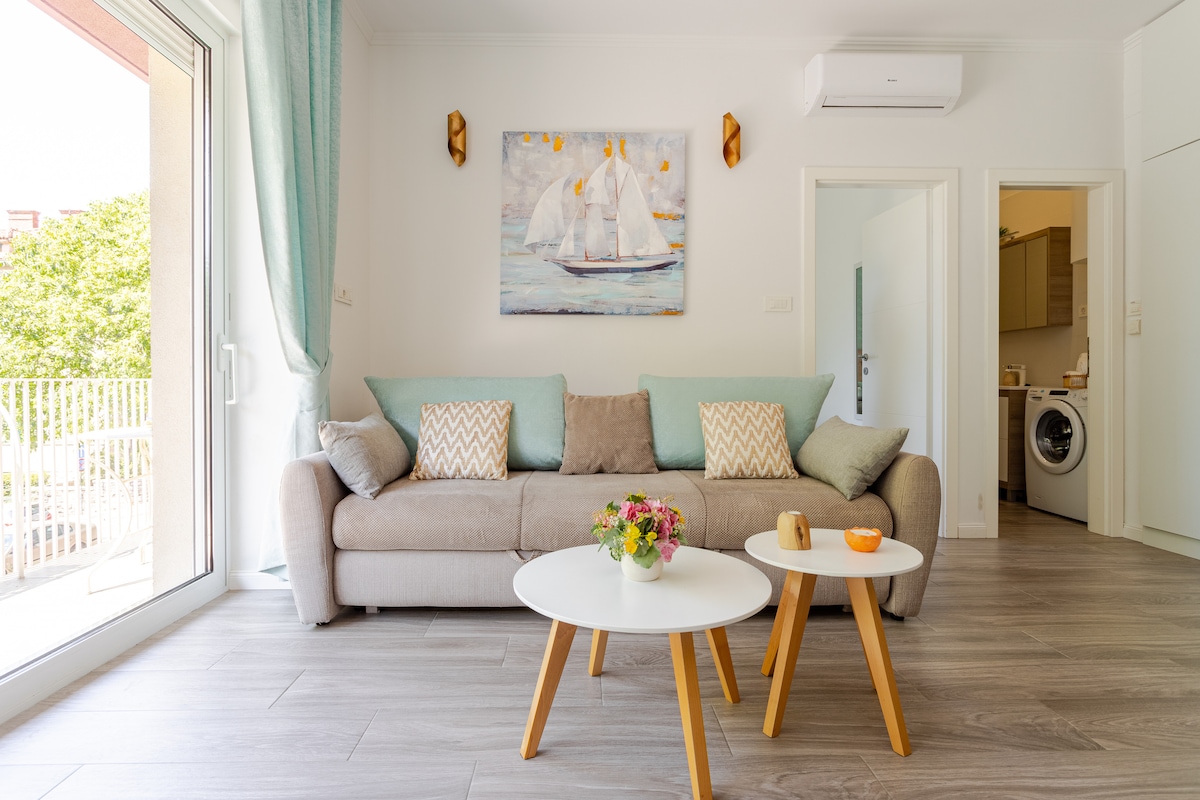 A comfortable living area is displayed, featuring a soft, light-colored sofa adorned with decorative cushions. A white coffee table sits in front, showcasing a small flower arrangement and an orange. Natural light floods the space through large glass doors, leading to a balcony.