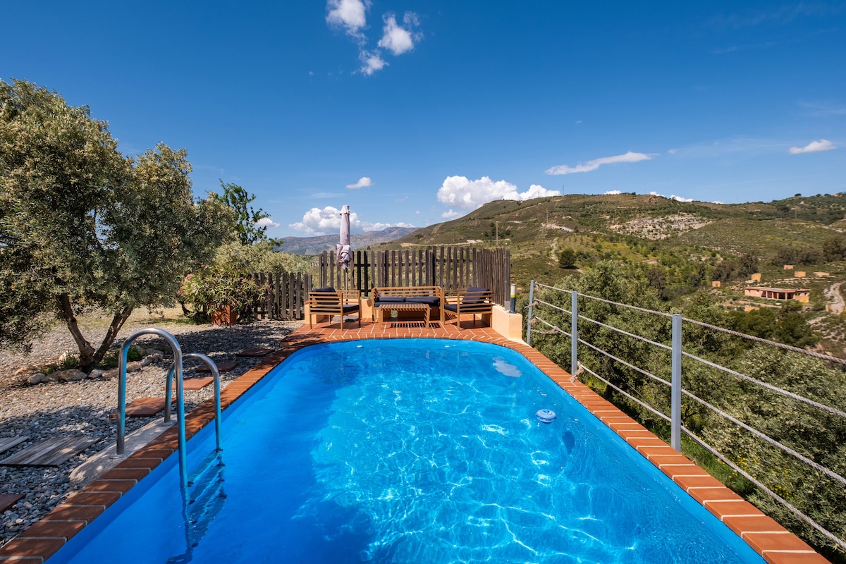 A tranquil swimming pool is featured, surrounded by wooden decking and lounge chairs. Scenic mountain views provide a breathtaking backdrop, emphasizing the property’s connection to nature under a clear blue sky with a few scattered clouds.