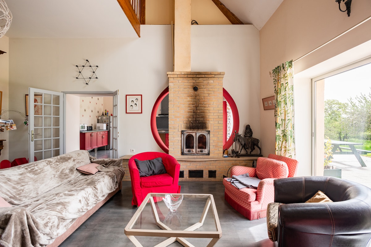 A spacious living area features a cozy fireplace made of brick, with a large, light-colored sofa draped in a soft throw. Comfortable red chairs are arranged around a glass coffee table. Large sliding doors lead to the outdoor terrace, allowing natural light to fill the space.