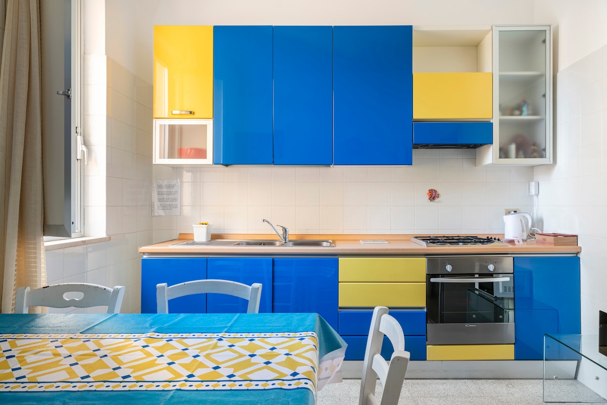 A modern kitchen is displayed with bright blue and yellow cabinetry. The space features a gas stove, oven, and sink, along with a dining area highlighted by a blue tablecloth. Natural light is allowed in through the window, enhancing the vibrant colors.