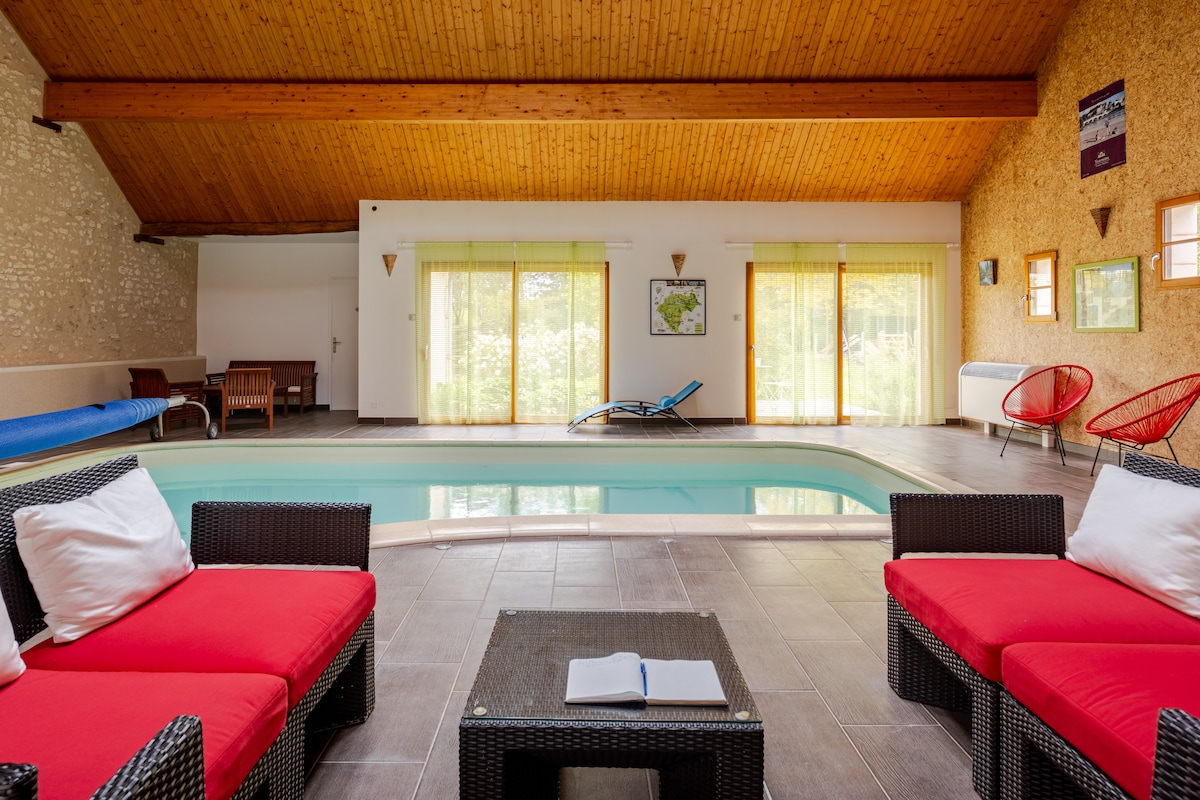 An indoor heated pool area features large glass doors that open to a view of surrounding greenery. Comfortable seating is arranged around a low table, with bright red cushions contrasting against the dark wicker. Natural wood accents on the ceiling enhance the room's warm ambiance.