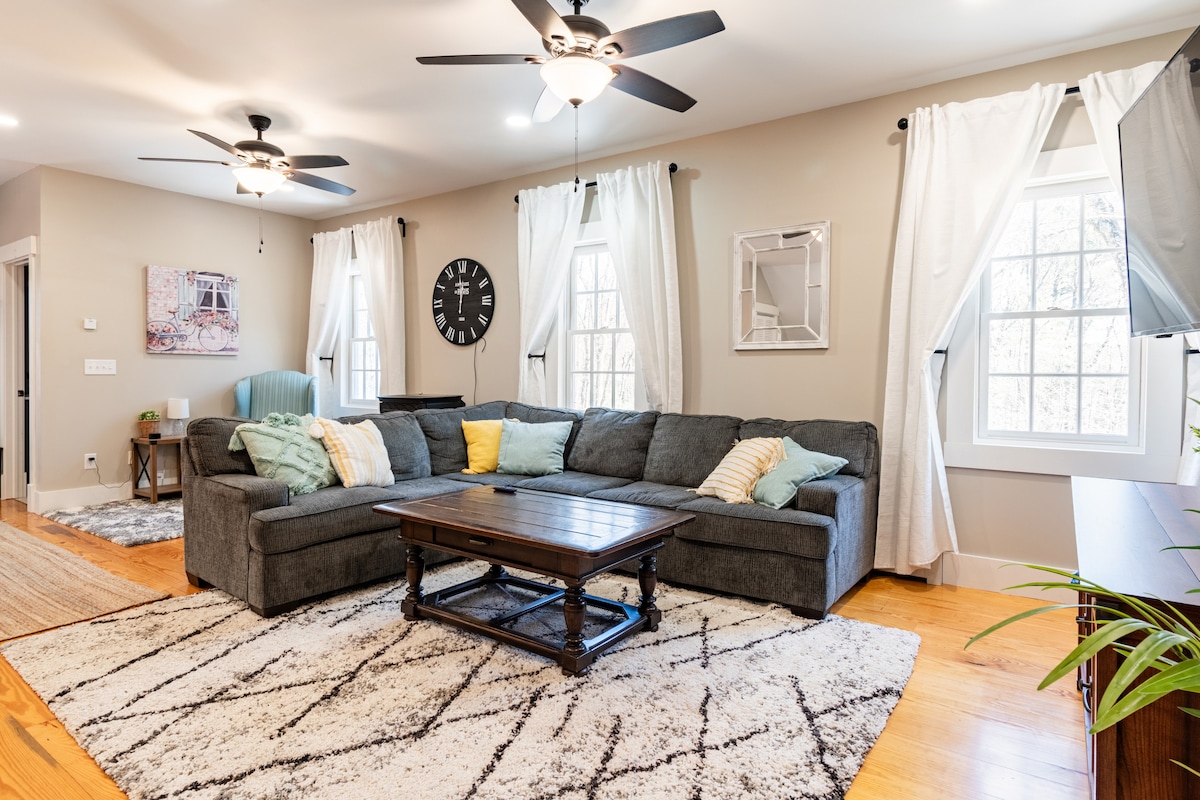 A spacious living area is furnished with a gray sectional sofa, accented with colorful throw pillows. A wooden coffee table is placed in front, on a soft area rug. Two large windows allow natural light to brighten the room, complemented by ceiling fans and tasteful decor.
