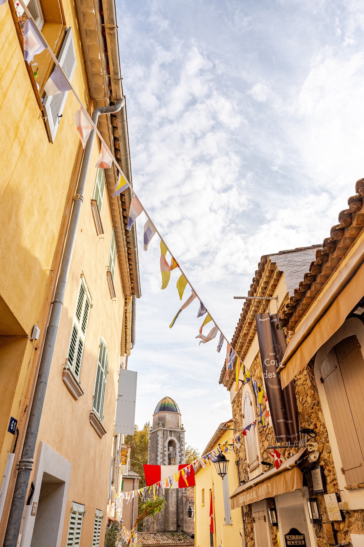 Colorful flags adorn a charming street in the historic center of Saint Tropez. Soft sunlight casts gentle shadows on the pastel-colored buildings, while a bell tower is visible in the background, contributing to the village's quaint atmosphere.