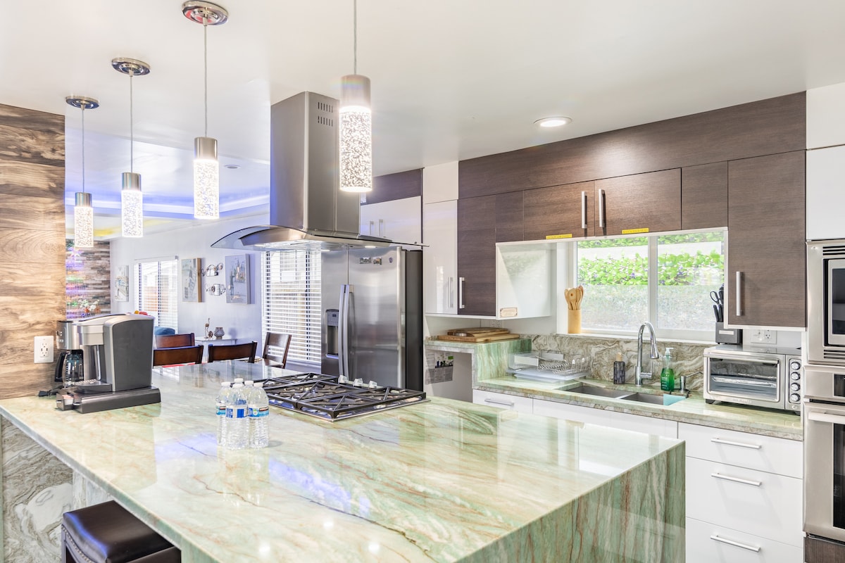 The modern kitchen showcases a large green marble countertop paired with sleek cabinetry. Pendant lights hang above, illuminating the space. Stainless steel appliances, including a refrigerator and oven, are visible. A window provides natural light, with kitchen utensils neatly arranged nearby.