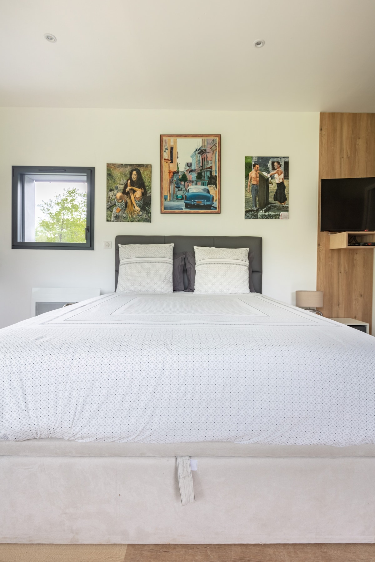 The sleeping area features a spacious bed dressed in white linens, flanked by two bedside lamps. Above the bed, three framed art pieces add character to the neutral-toned walls, with a window allowing natural light to illuminate the space.
