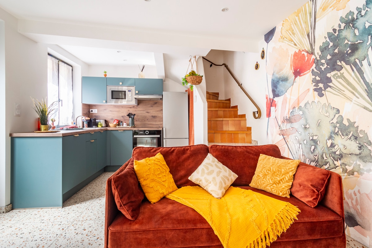The living area showcases a cozy sofa adorned with decorative cushions and a vibrant throw blanket. A well-equipped kitchen is visible in the background, featuring modern appliances and a staircase leading to the upper level, enhancing the space's overall functionality.