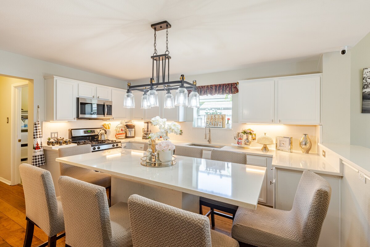 A modern kitchen is featured, showcasing white cabinetry and a large island with seating for several guests. Overhead, a statement chandelier with multiple glass pendant lights provides illumination. A stove and stainless steel appliances are present, alongside decorative elements that add warmth to the space.