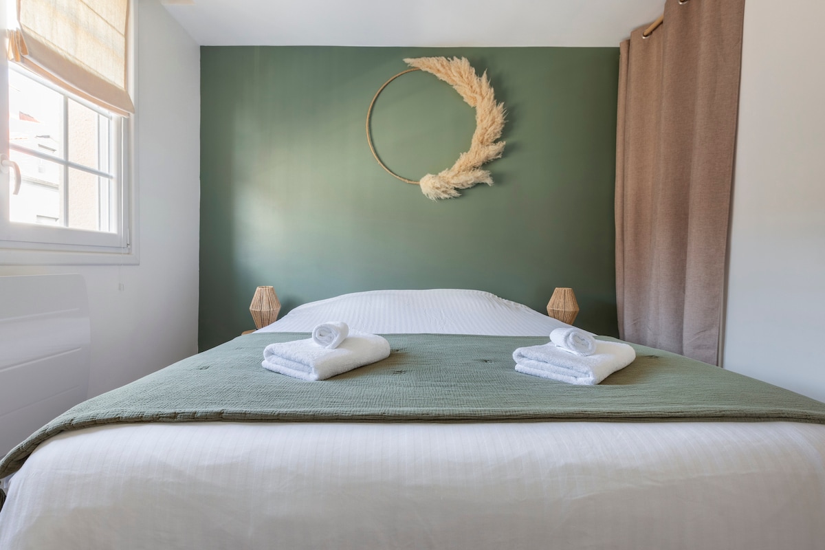 A calm bedroom features a spacious bed dressed in crisp white linens and a soft green throw. Two neatly rolled towels are placed on the bed. A circular wall decoration complements the muted green backdrop, while natural light filters through a window covered by a light shade.