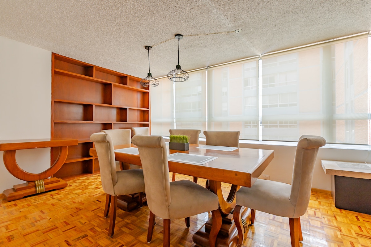 A dining area features a wooden table surrounded by six beige upholstered chairs. Natural light floods the space through large windows, complemented by a minimalist bookshelf against the far wall. The flooring is made of polished wood, creating a warm ambiance.