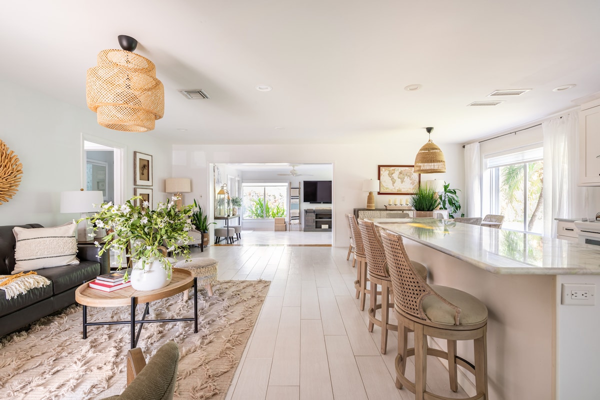 A spacious living area features a comfortable sofa and a large circular coffee table centered on a soft area rug. Natural light filters through multiple sliding doors that provide views of the outdoor space. A well-appointed kitchen with bar seating is visible in the background.