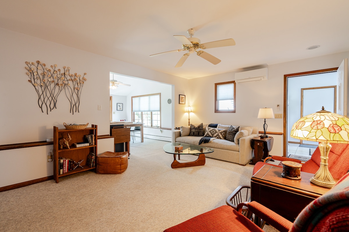 A spacious living area features a light carpeted floor and neutral walls. A comfortable sofa is positioned alongside a glass coffee table, with a ceiling fan above for airflow. Warm lighting from a lampshade complements a wooden bookshelf and additional seating.