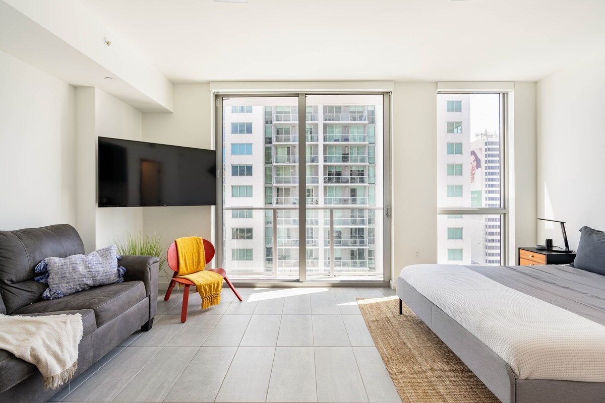 A spacious room features large windows providing ample natural light and city views. A sleek television is mounted on the wall opposite a relaxation area with a gray sofa and a colorful accent chair. A neutral-toned bed is positioned alongside a natural fiber rug.