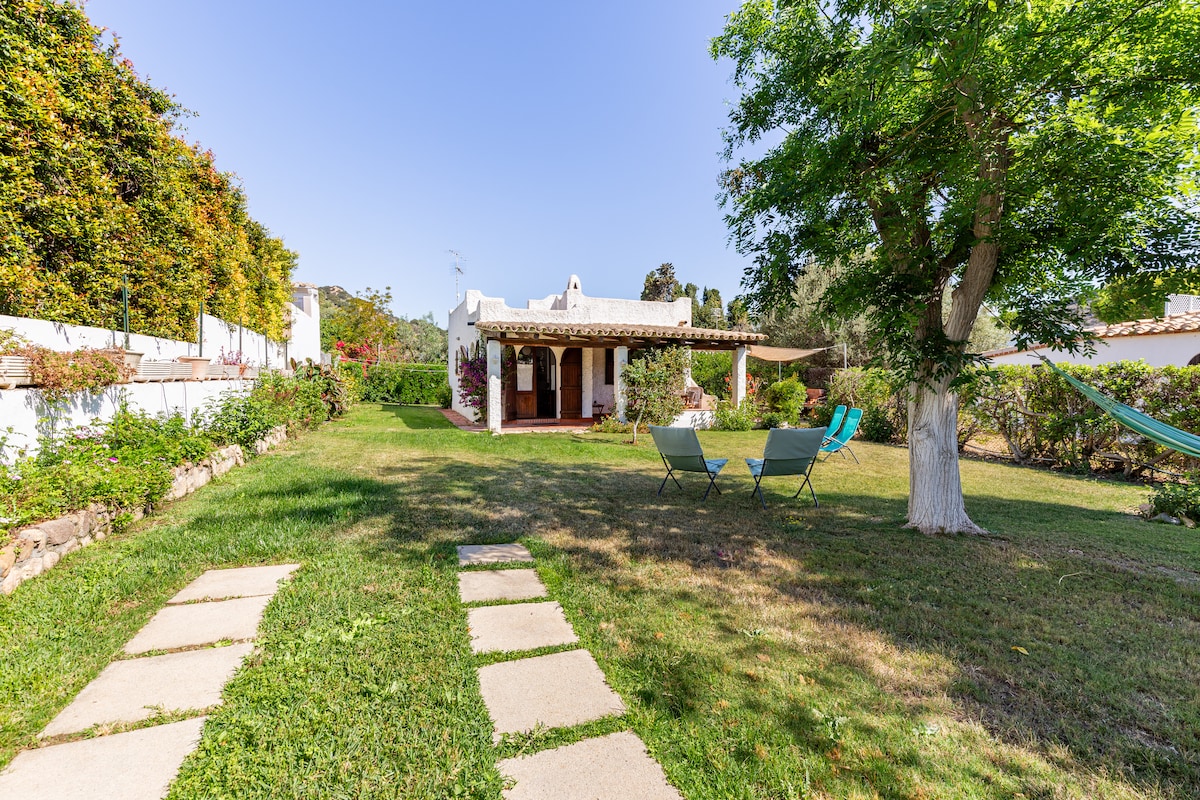 The spacious garden features a lush green lawn bordered by flower beds and pathways. A cozy seating area with two chairs is positioned on the grass, while a hammock hangs invitingly near a tree. The villa is visible in the background, surrounded by greenery.