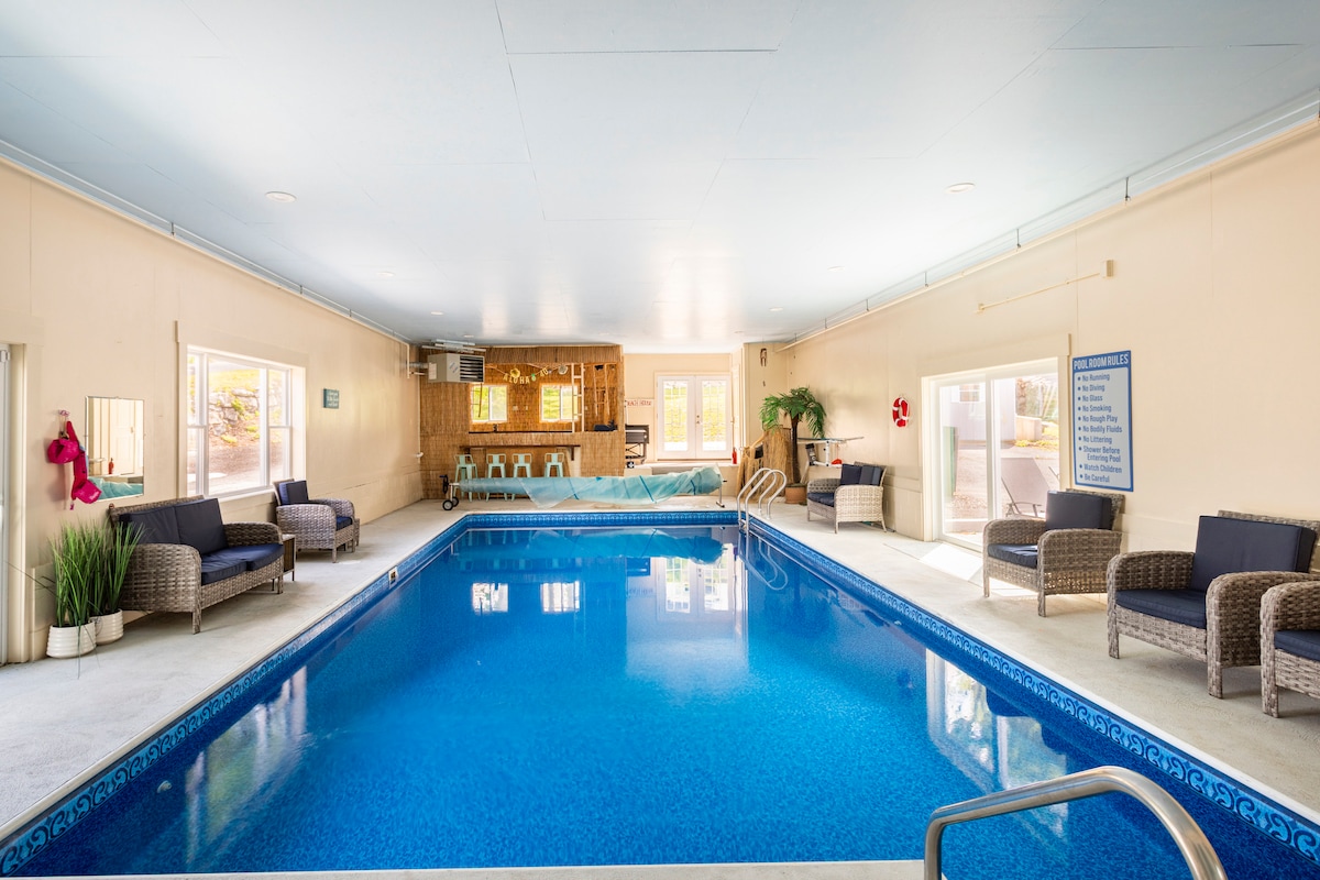 An indoor heated pool area features crystal-clear blue water, surrounded by comfortable lounge seating. Natural light enters through the windows, creating a warm ambiance. A bar area is positioned nearby, with a safety sign visible on the wall. The space is designed for relaxation and enjoyment year-round.